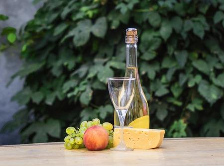 Composition of cheese, grapes, peaches, white grapes, bottles and glasses of wine on a wooden round table. in the courtyard. against the background of the vineの写真素材