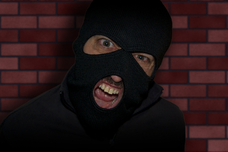 man close up thief in a mask and a blue shirt on a yellow background looks slyly to the camera. Mimicry. Photo shoot. Evil criminal wearing balaclavaの写真素材