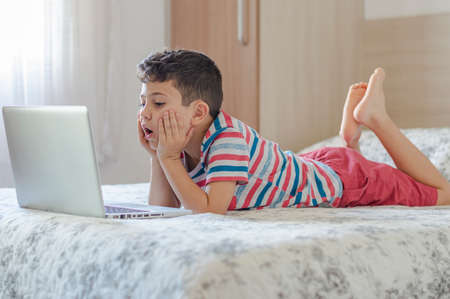 Young boy child lying on bed using a laptop. Online class lesson on video. Distant remote education. New normal school study. Kids at schoolの写真素材