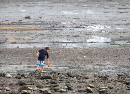 KRABI,THAILAND-AUGUST 18, 2018 : The boy is looking for something on the beach filled with rubbish,reef and dirty mud.のeditorial素材