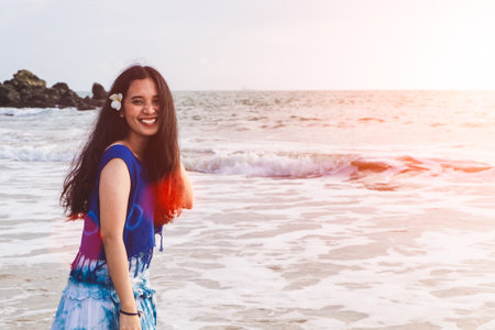 Asian girl wear Indigo dyed cotton long skirt,smiling and flower tucked behind her ear on the beach,blue sky and sea,orange light flare.の写真素材