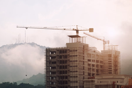 Tall buildings that are being built in the middle of a small town Close to the mountains With cranes are working The background is cloudy and the sunlight is mild in the morning. Under the concept of a growing cityの写真素材