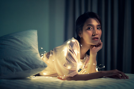 A young woman lying and playing with sparkling lights or fairy light on a white bed In a room with a dim yellow light.の写真素材