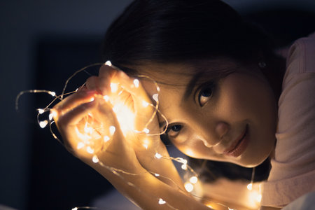 A young woman lying and playing with sparkling lights or fairy light on a white bed In a room with a dim yellow light.の写真素材