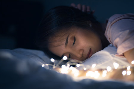 A young woman lying and playing with sparkling lights or fairy light on a white bed In a room with a dim yellow light. She is closing her eyes.の写真素材