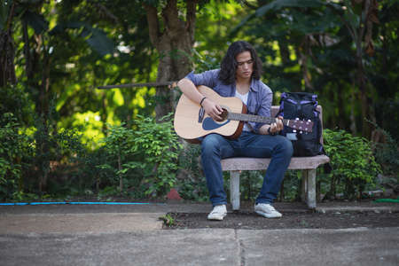 Asian long haired man is playing the guitar while he is traveling. With a rustic background and trees Under the soft sunlight in the dayの写真素材