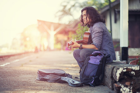 Asian long haired man is playing the guitar while he is traveling. With a rustic background and trees Under the soft sunlight in the dayの写真素材