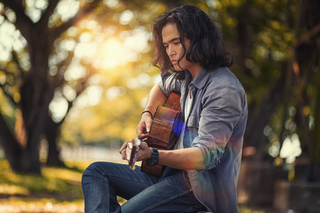 Asian long haired man is playing the guitar while he is traveling. With a rustic background and trees Under the soft sunlight in the dayの写真素材