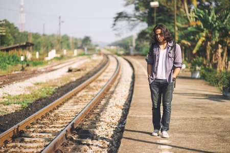 Long haired man that looks cool He is a traveler or nomadic musician who is backpacking and guitars, walking near train track in rural.の写真素材