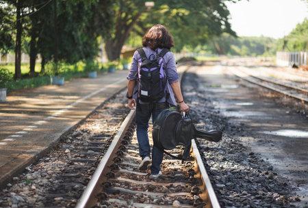 Long haired man that looks cool He is a traveler or nomadic musician who is backpacking and guitars, walking near train track in rural.の写真素材