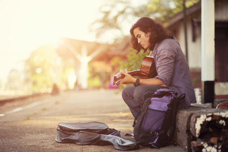 Asian long haired man is playing the guitar while he is traveling. With a rustic background and trees Under the soft sunlight in the dayの写真素材