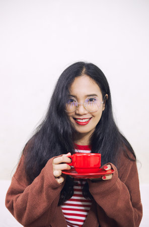Asian women are holding a cup of red coffee. She wore a white-red striped shirt and a brown sweater. Apply red lipstick and red nail polish. She sat in front of an isolated white background.の写真素材