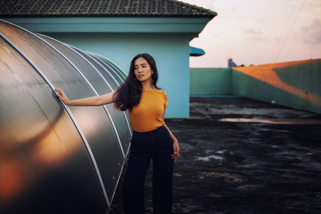 The girl has a skin color of honey or tan. Wearing orange shirt Street photography poses on rooftop and orange lightの写真素材