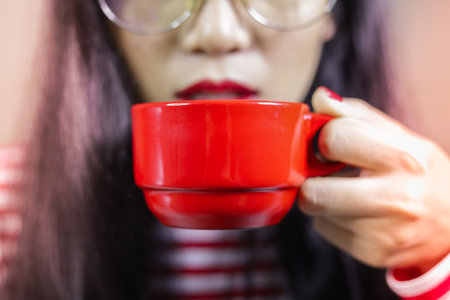 Asian women wearing glasses are holding red coffee mugs She wore long-sleeved striped shirts with white-red stripes. She painted a red nail, a depth of field close-up, with red light, light orange.の写真素材