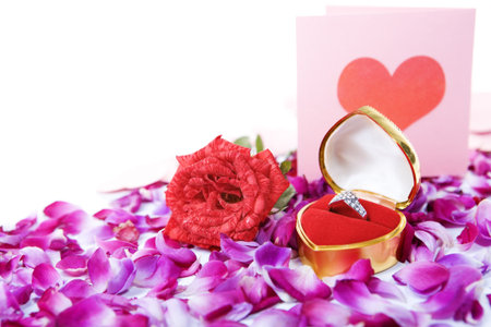 Ring in container with single rose and a valentine card paced together above rose petal with with...