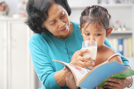 Grandma giving milk to her granddaugter while she busy reading bookの写真素材