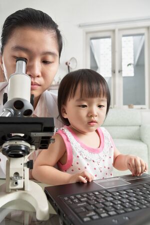 Mother busy working with microscope and her daughter busy playing laptopの写真素材