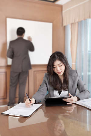 Chinese businesswoman busy working with her tablet while businessman on background busy preparing presentationの写真素材