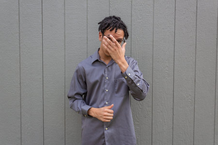 Biracial man looking between fingers of hand over face, adjusting eyeglasses, natural hair in...