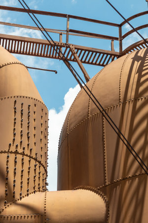 Heavily riveted metal tanks and catwalk, industrial shapes against a blue sky with clouds, vertical aspectの写真素材