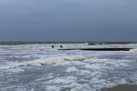 Distant surfers in a storm, heavy surf and sea foam on a sandy coastline, horizontal aspect