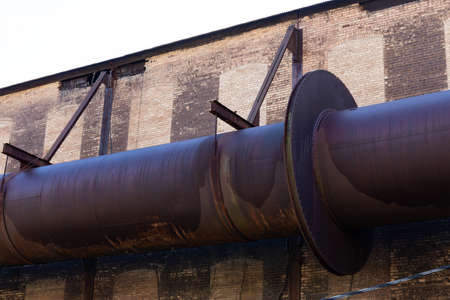 Enormous industrial pipe running along the exterior of a brick manufacturing warehouse, rusted metal and huge flanges riveted together, horizontal aspectの写真素材