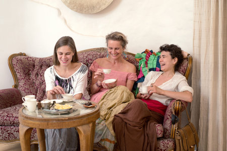 Three women in ethnic dresses sitting on an old couch and drinking teaの写真素材