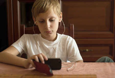 Boy is sitting at the table and looking at the smartphoneの写真素材