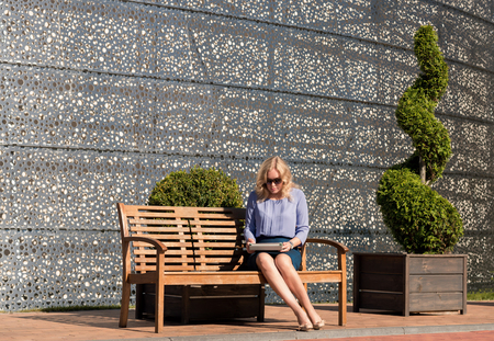 Blonde woman is sitting on a bench with a digital tabletの写真素材