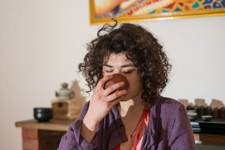 Young Asian woman with curly hair drinking tea made of clay mug