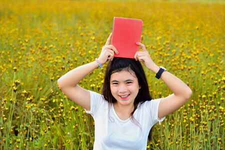 Cute Asian girl holds red book on head. In the middle of a yellow flower field, thoughts, love, reading and education.の写真素材