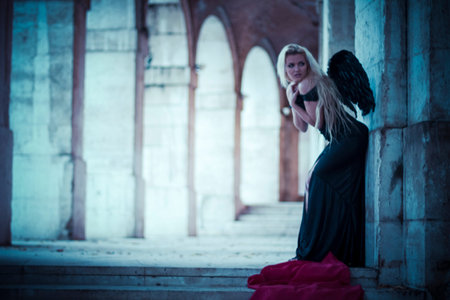 Fallen, an angel in the ruins of a city, beautiful blonde with long hair dressed in black and with black wingsの写真素材