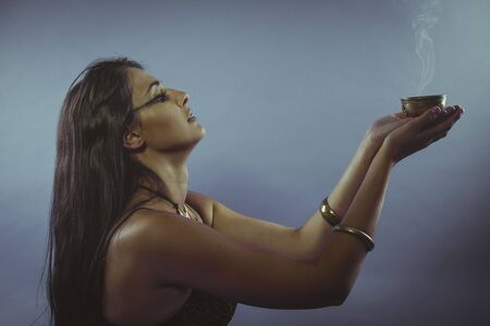 Witchcraft, Tribute and prayer. Beautiful brunette with bronze bowl and incense in a gesture of faithの写真素材