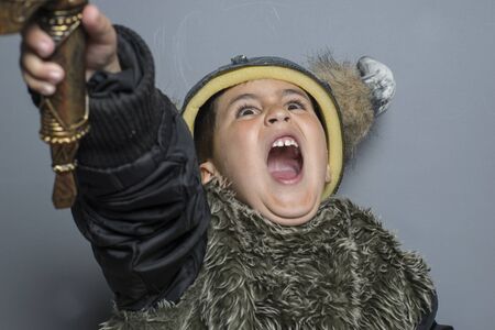viking brunette boy in fur shirt in preschool age standing indoors, wearing costume hat with horns, hands up, making a scary face and swordの写真素材