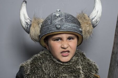 viking brunette boy in fur shirt in preschool age standing indoors, wearing costume hat with horns, hands up, making a scary face and swordの写真素材