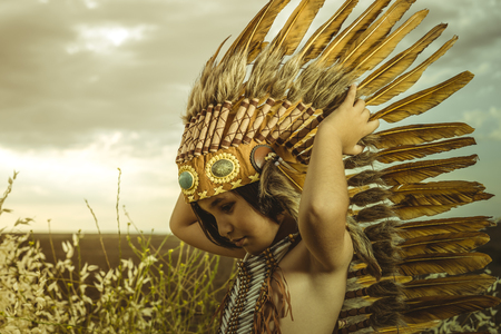 Child dressed as an American Indian at sunset, wearing an Indian feather plume and breastplate of bones. Playing next to a cereal meadowの写真素材
