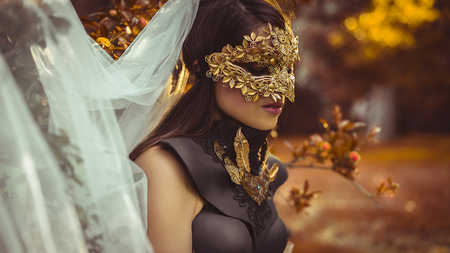 Golden, mysterious woman dressed in white with mask of leaves in copper and gold color in a garden in autumnの写真素材