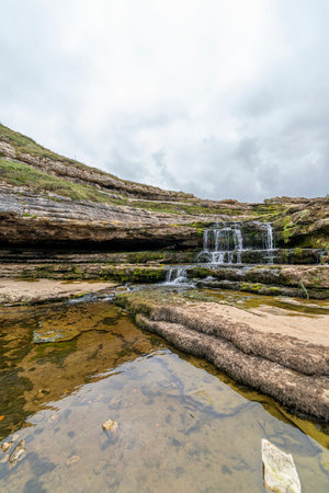 Picturesque waterfall flowing over layered rock formations with lush green moss in Cantabria, Spain. Ideal for nature, travel, and landscape themes.の写真素材