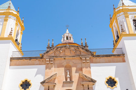 The stunning facade of the Church of Nuestra SeÃ±ora de la Merced in Ronda, Spain, featuring twin bell towers and a central fountain under a bright blue sky. Perfect for themes of Spanish architecture, religious landmarks, and cultural tourismの写真素材