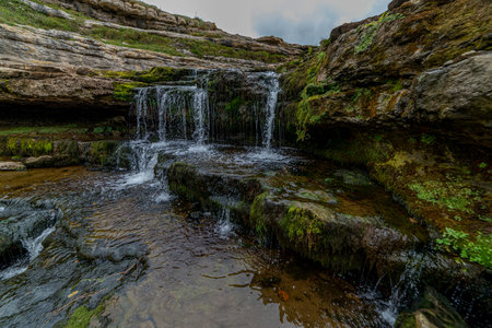 Picturesque waterfall flowing over layered rock formations with lush green moss in Cantabria, Spain. Ideal for nature, travel, and landscape themes.の写真素材