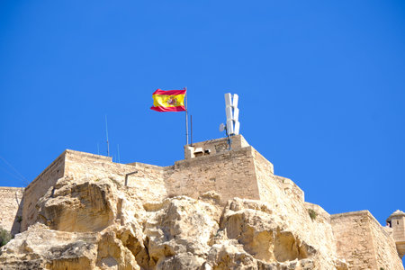Spanish flag waves on historic castle against clear blue sky.の写真素材