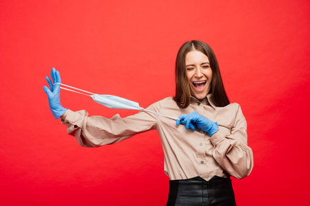 Cheerful and joyful young female playing with protective medical mask and blue gloves on red...