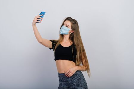 Long haired girl with surgical mask looking at the smartphone and doing physical exercise. Sport in quarantine concept.の写真素材