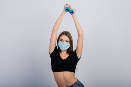 Young woman in a protective mask with dumbbells on a white background. Quarantined sportsの写真素材