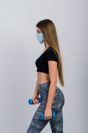 Young woman in a protective mask with dumbbells on a white background. Quarantined sportsの写真素材