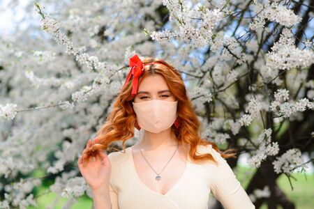 Close up portrait of tender girl in a white blouse under a blossoming cherry tree with a mask with flowers on from the coronavirus.の写真素材