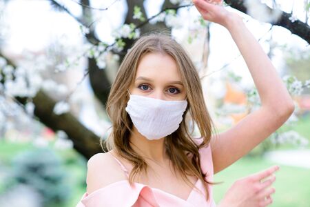 Close up portrait of tender girl in a white blouse under a blossoming cherry tree with a mask with flowers on from the coronavirus.の写真素材