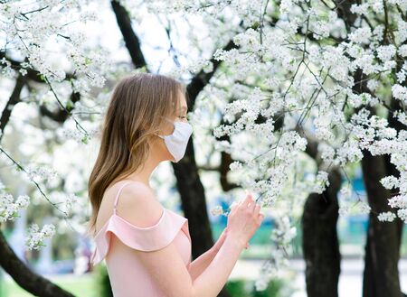 Close up portrait of tender girl in a white blouse under a blossoming cherry tree with a mask with flowers on from the coronavirus.の写真素材