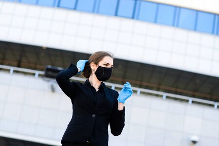 Beautiful businesswoman in a black suit in a black medical mask and gloves at the city in quarantine and isolation. Pandemic COVID-19. Image with selective focusの写真素材