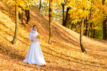 Cute girl in long white wedding dress posing in rural path among autumnal trees in forest in golden hour atmosphere.の写真素材